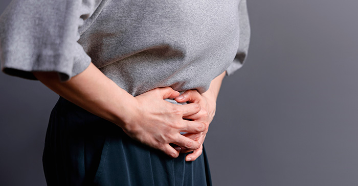 A imagem ilustra uma mulher de blusa de frio cinza e calça azul escura com as duas mãos na barriga e meio inclinada, como se estivesse sentindo dor